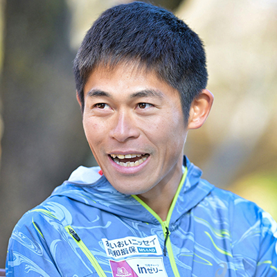 男性が青いジャケットを着て笑顔で話している。背景はぼけている。