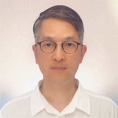 眼鏡をかけた男性が白いシャツを着て、カメラを見つめている。背景は淡い色合い。