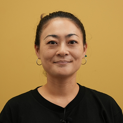 女性が黄色い背景の前で微笑んでいます。黒いシャツを着用し、シンプルな印象です。