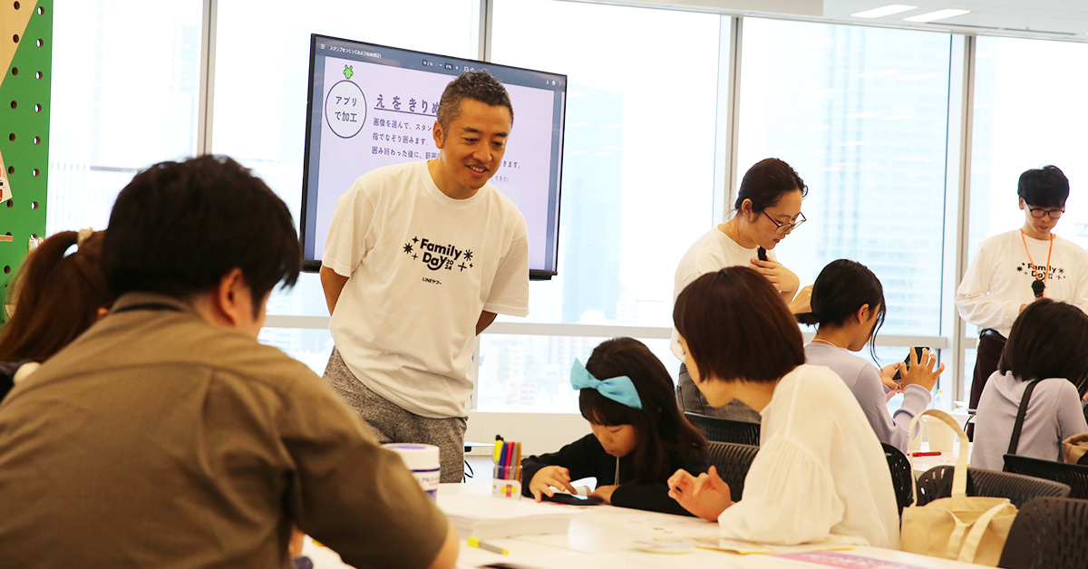 家族連れがテーブルで楽しそうに活動しています。背景には大きなスクリーンがあります。