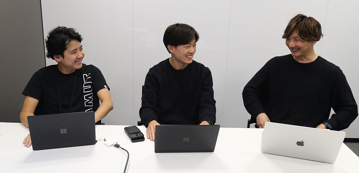 オフィスの会議テーブルでラップトップを前に笑顔で取材に応じる3名（取材風景）