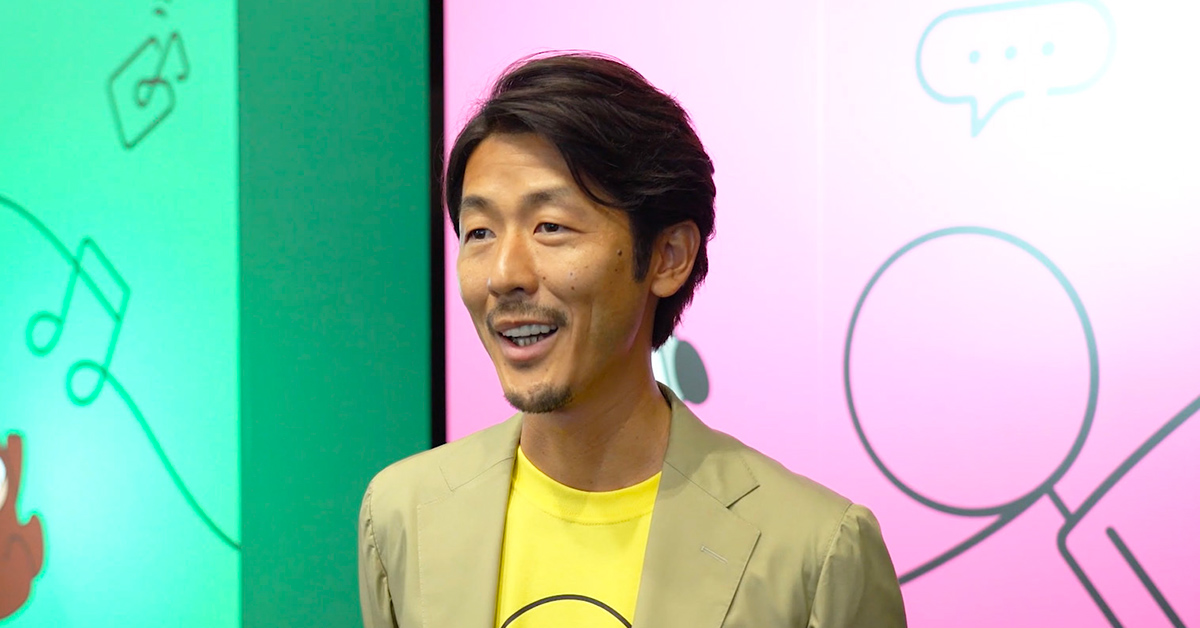男性がカラフルな背景の前で話しています。イベントでのスピーチの一場面を切り取った写真です。