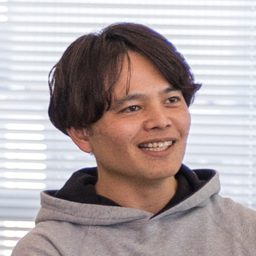 画像には、フード付きのパーカーを着た男性が笑顔で写っています。背景にはブラインドのある窓が見えます。