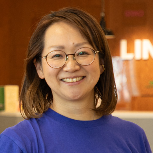 眼鏡をかけた女性が、木目調の背景の前で笑顔を見せています。