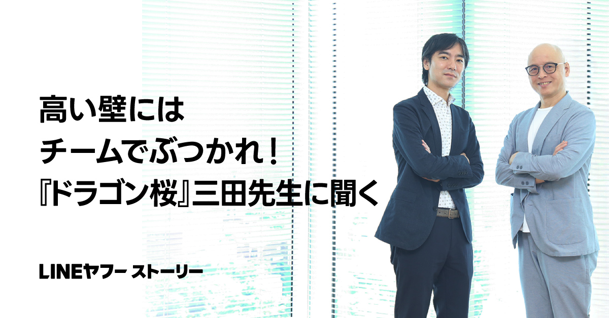 二人の男性がスーツを着て立っています。背景には「高い壁にはチームでぶつかれ！」というテキストがあります。