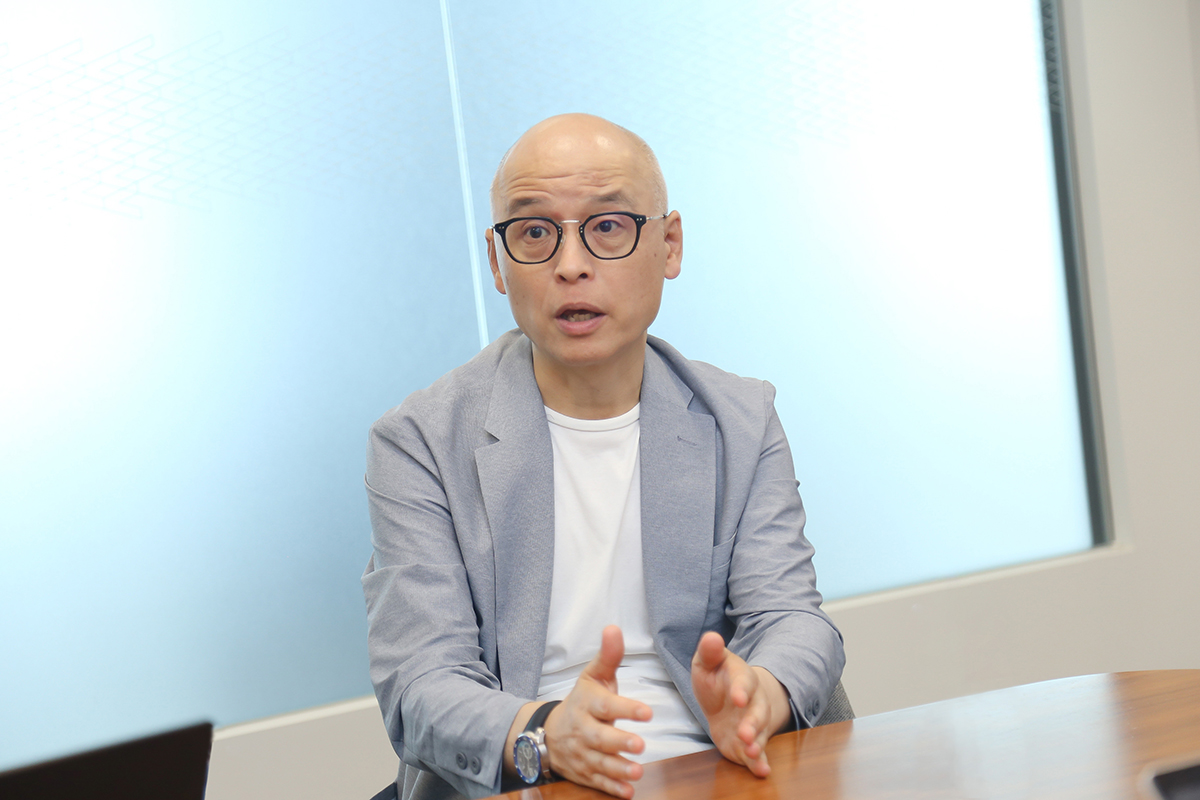 スーツを着て眼鏡をかけた男性がテーブルに座り、手を使って話をしています。背景はシンプルな室内です。