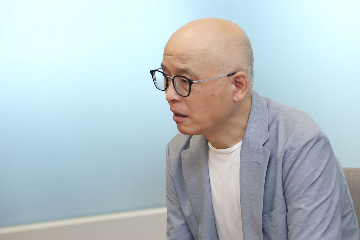 スーツを着た男性が、横を向いて微笑んでいます。背景はシンプルな室内で、落ち着いた雰囲気です。