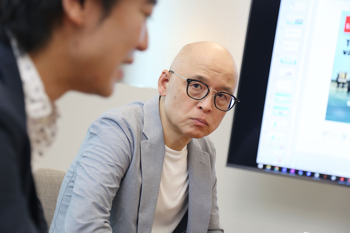 スーツを着て眼鏡をかけた男性が、座って他の人の話を聞いている様子です。背景にはスクリーンが見えます。