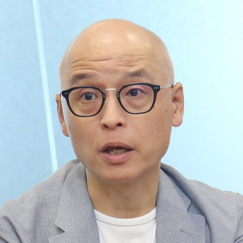 スーツを着て眼鏡をかけた男性が、正面を見て話をしています。背景はシンプルな室内です。