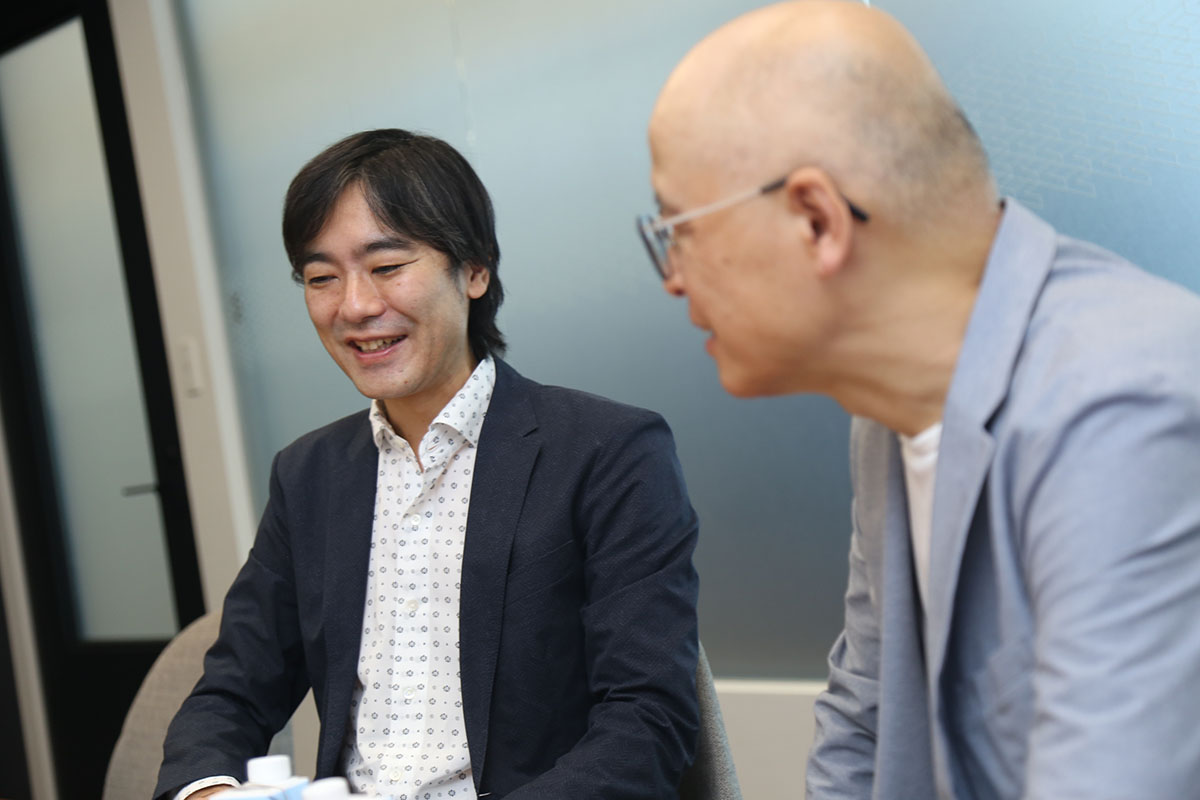 スーツを着た男性が笑顔で話をしています。背景はシンプルな室内で、和やかな雰囲気です。