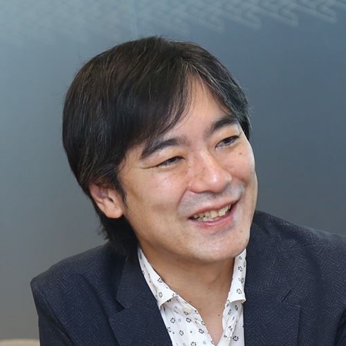 スーツを着た男性が、横を向いて微笑んでいます。背景はシンプルな室内で、落ち着いた雰囲気です。