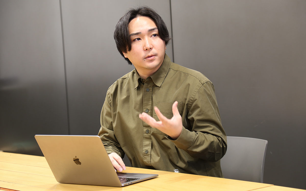 カジュアルなシャツの男性がノートパソコンを前に話している。