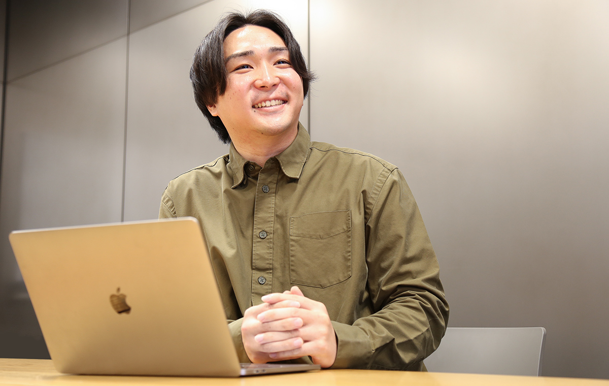 カジュアルなシャツの男性がノートパソコンを前に笑顔で座っている。