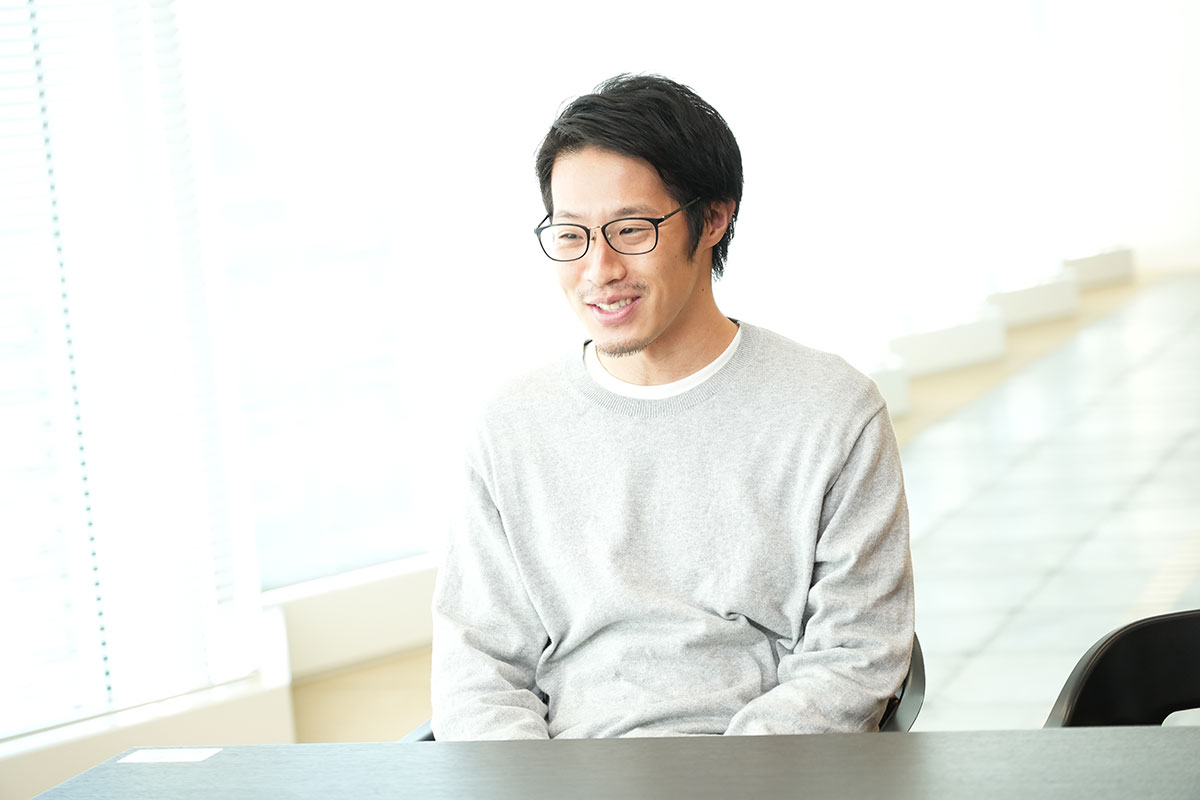 明るいオフィスで眼鏡をかけた男性が座っている写真です。彼はリラックスした様子で微笑んでいます。