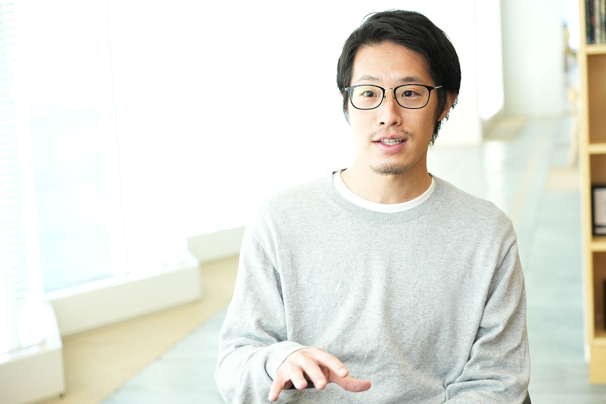 明るいオフィスで眼鏡をかけた男性が座っている写真です。彼はリラックスした様子で微笑んでいます。
