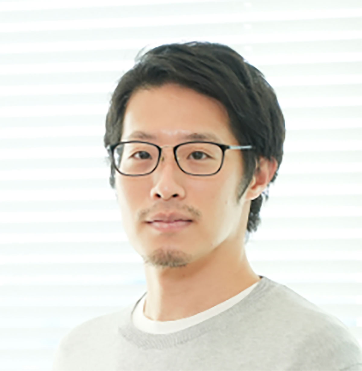 明るいオフィスで眼鏡をかけた男性が座っている写真です。彼はリラックスした様子で微笑んでいます。