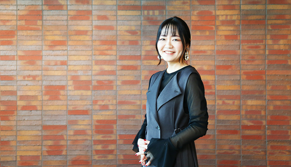 レンガの壁の前で黒い服を着た女性が笑顔で立っている。
