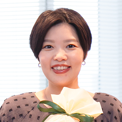 女性が室内で微笑み、プレゼントを持っている。背景のブラインドが光を通し、明るい雰囲気が漂う。