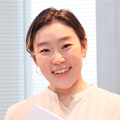 女性が室内で微笑み、白い服を着ている。背景のブラインドが光を通し、明るい雰囲気が漂う。