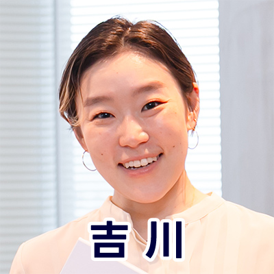 女性が室内で微笑み、白い服を着ている。背景のブラインドが光を通し、明るい雰囲気が漂う。名前が「吉川」と表示されている。