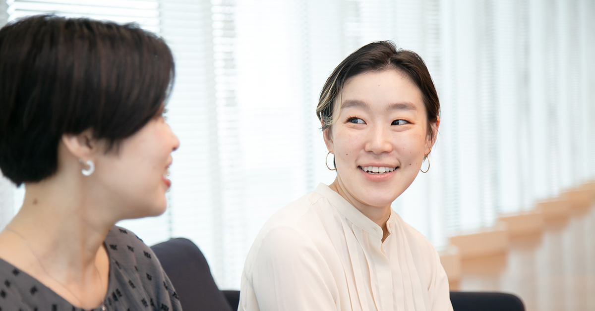 二人の女性が室内で談笑している。背景のブラインドから光が差し込み、明るい雰囲気が漂う。