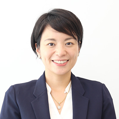 女性が笑顔でカメラを見つめている。白い背景に、ビジネススーツを着用。