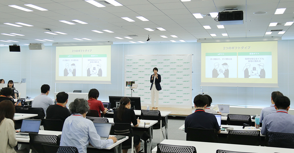 プレゼンテーションを行う女性がステージに立ち、聴衆が座っている。スクリーンにはスライドが表示。