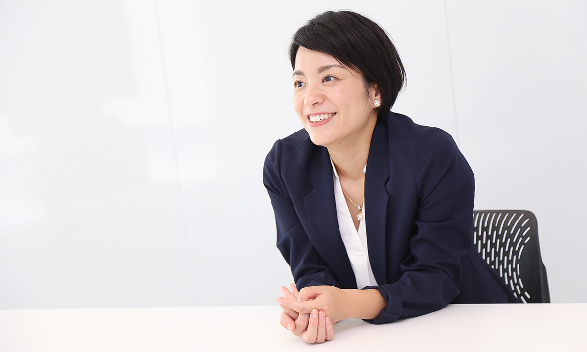 女性がテーブルに肘をつき、笑顔で話している。明るい室内でリラックスした雰囲気。