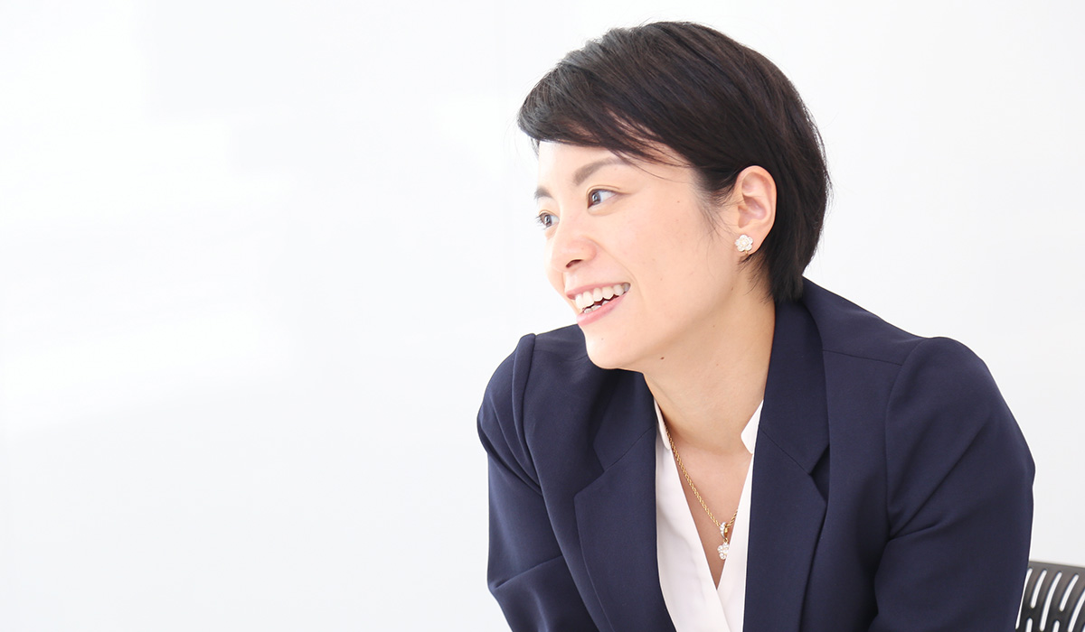 女性が横を向いて笑顔で話している。白い背景に、ビジネスカジュアルな服装。