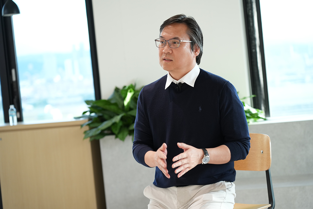 眼鏡をかけた男性が、椅子に座って話している様子です。背景には窓と観葉植物があり、オフィスの一角のようです。