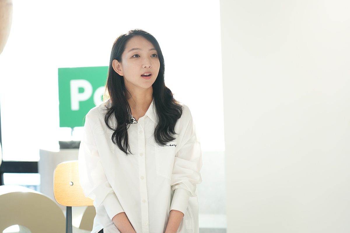 白いシャツを着た女性が椅子に座って話している様子です。背景に「Pay」の文字が見え、明るい室内での撮影です。