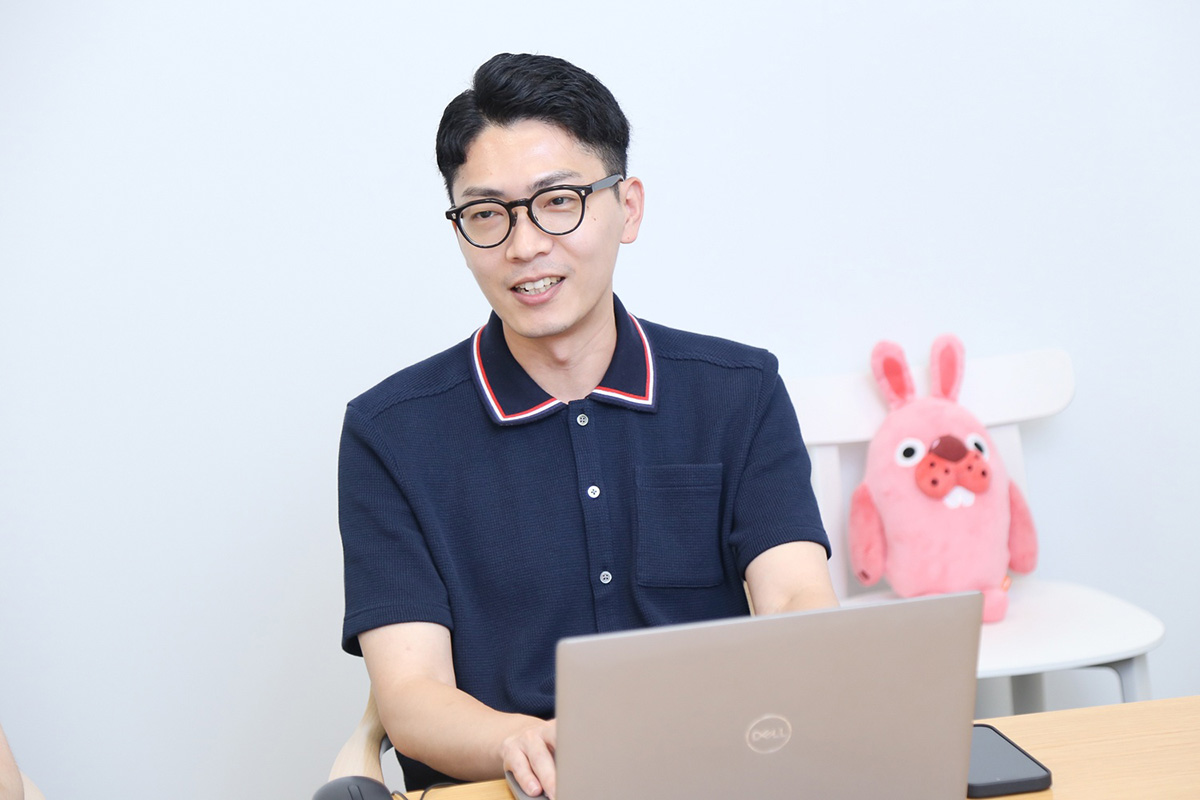 眼鏡をかけた男性がノートパソコンを前にして話している。後ろにはピンクのぬいぐるみ。