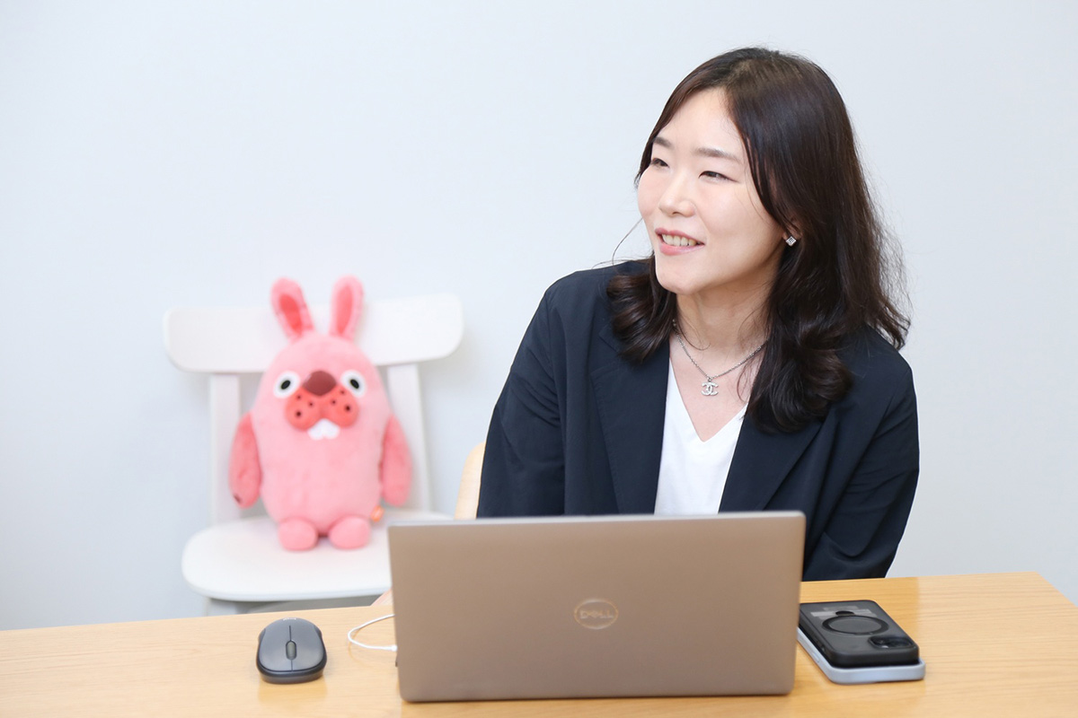 女性がノートパソコンを前にして笑顔で話している。後ろにはピンクのぬいぐるみ。