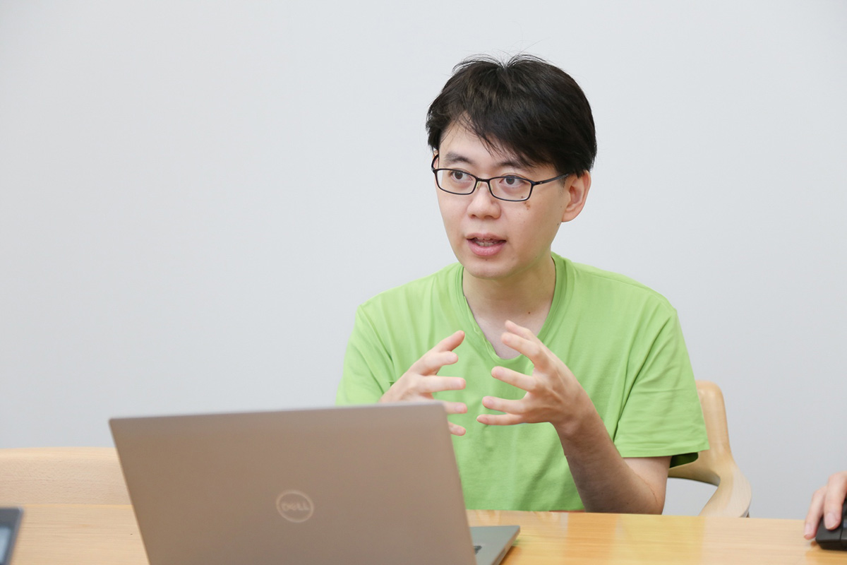 緑のシャツを着た男性がノートパソコンを前にして話している。真剣な表情。