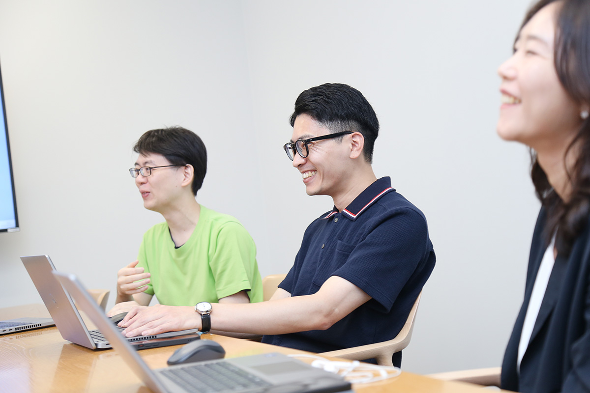 三人の人物が笑顔で座り、ノートパソコンを前にして会話している。