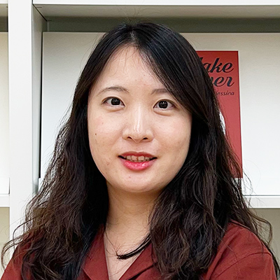 女性がカメラに向かって微笑んでいる。背景には棚があり、落ち着いた雰囲気が漂う。