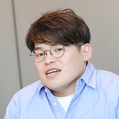 青いシャツを着た男性がカメラに向かって話している写真です。彼はメガネをかけており、リラックスした表情です。
