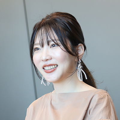 茶色のトップスを着た女性が微笑んでいる写真です。彼女は大きなイヤリングをつけ、明るい表情をしています。