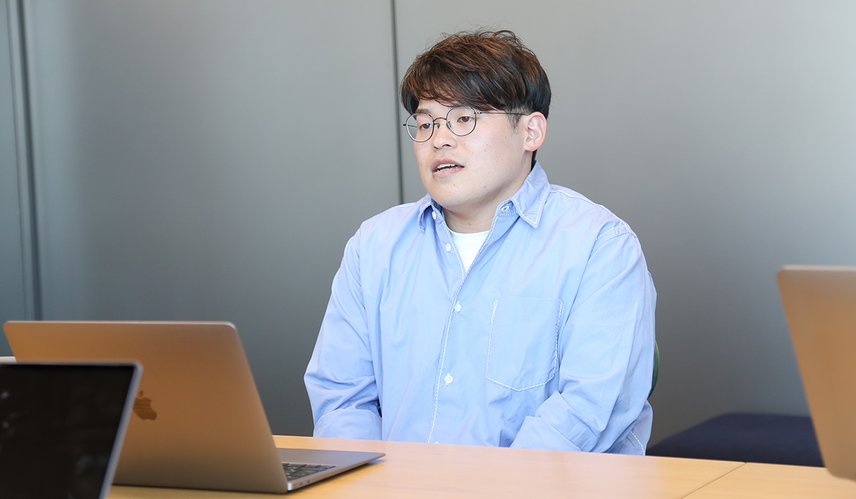 青いシャツを着た男性がラップトップの前で話している写真です。会議中の様子を捉えています。