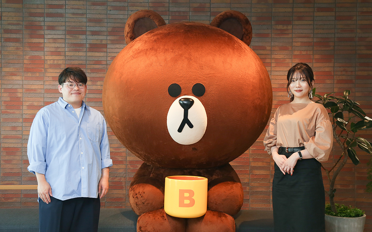 大きなブラウンのぬいぐるみと二人が並んでいる写真です。背景はレンガの壁で、親しみやすい雰囲気です。