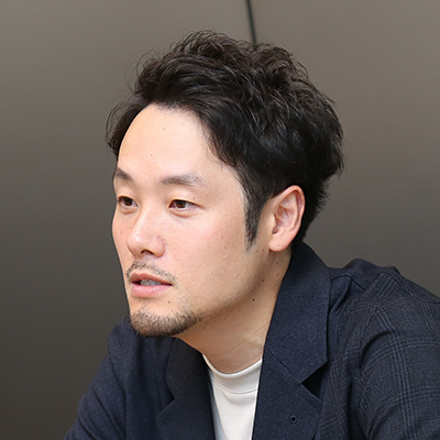 短髪の男性が真剣な表情で話している。ダークカラーのジャケットを着用し、落ち着いた雰囲気。
