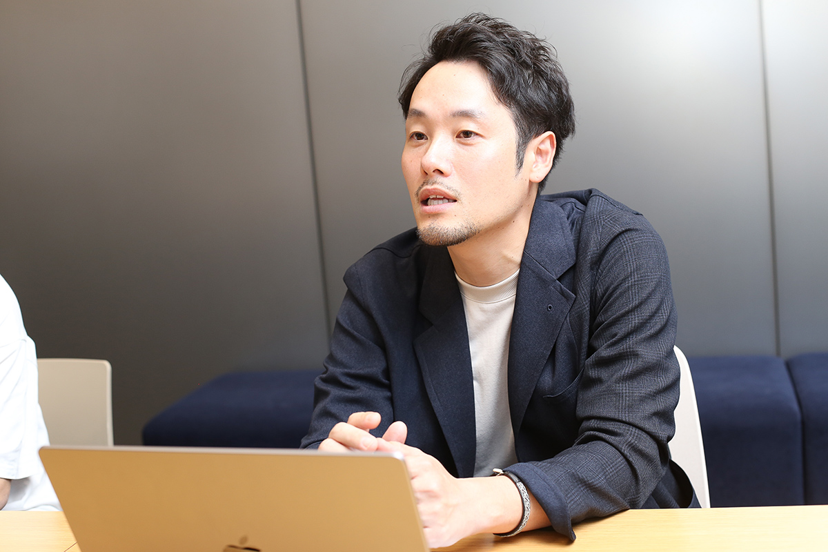 ダークカラーのジャケットを着た男性が会議中に話をしている。真剣な表情で、ノートパソコンを前にしている。