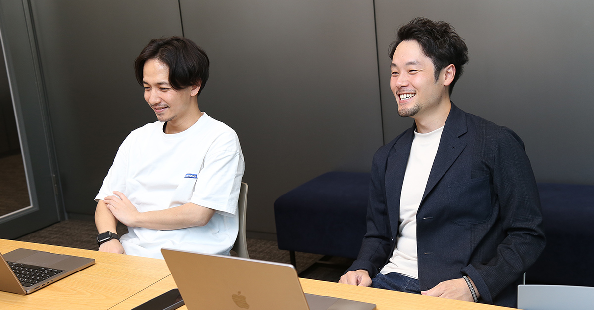 二人の男性が会議室で笑顔で座っている。リラックスした雰囲気で、ノートパソコンの前にいる。