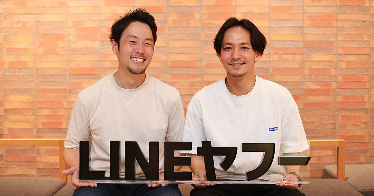 二人の男性が「LINEヤフー」のロゴを持って笑顔で座っている。レンガの背景が特徴的。