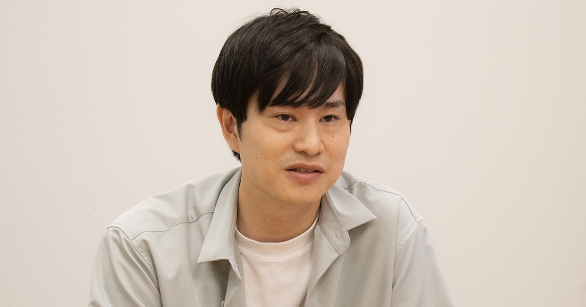 男性がインタビューに答えている様子。白い背景で、カジュアルな服装をしている。