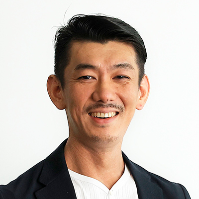 男性が白いシャツと黒いジャケットを着て笑顔でカメラを見ています。背景はシンプルな白色で、彼の表情が際立っています。