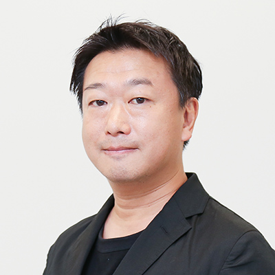 スーツ風の黒いジャケットを着た男性、小笠原が、白背景の前で穏やかに微笑んでいるポートレート写真です