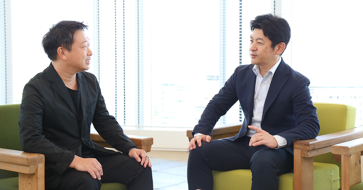 この画像には、明るいオフィスで男性2人、小笠原社長と田鎖社長が椅子に座って談笑している様子が写されています。リラックスした雰囲気の中でビジネスの会話が行われているようです