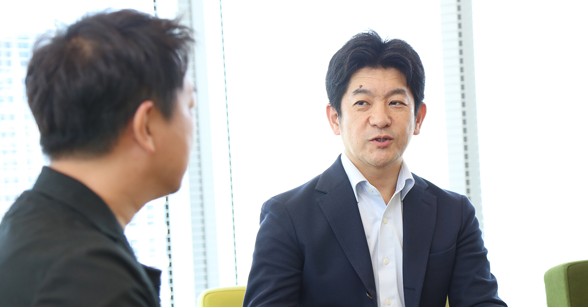 こちらは明るいオフィスで2人の男性経営者が対話している様子を写した写真です。右側の男性、小笠原が話し、左側の男性、田鎖が聞いています