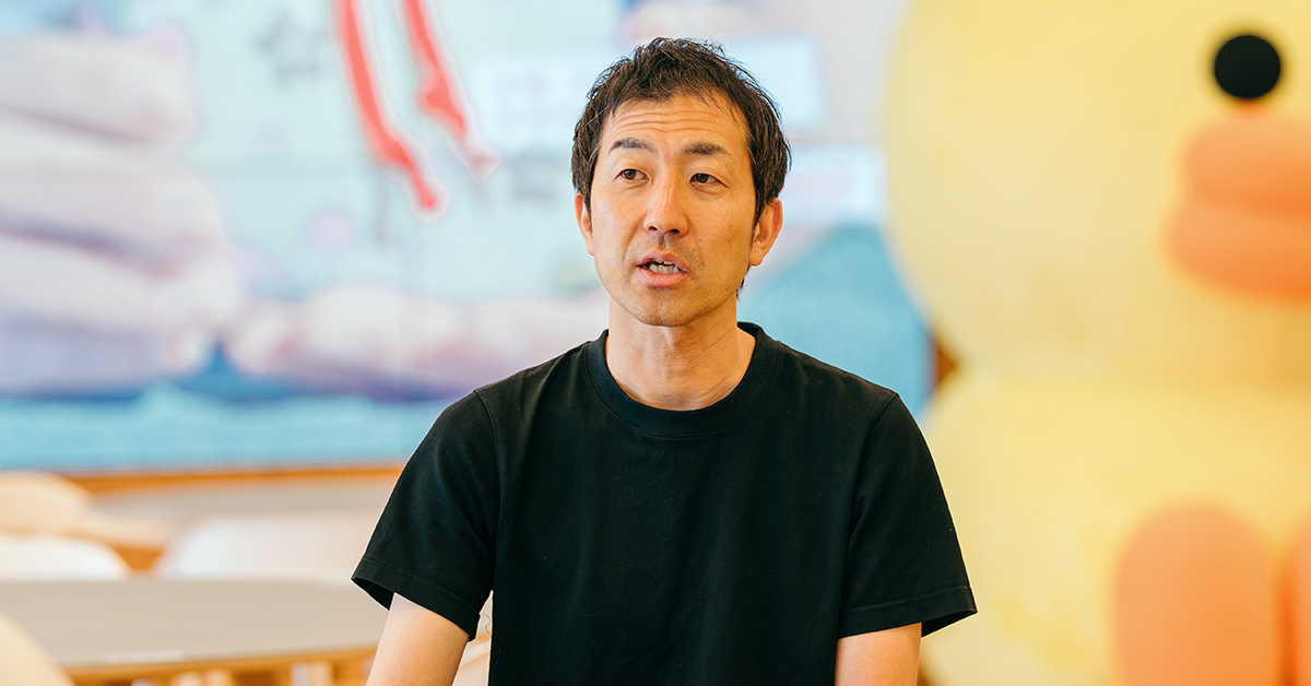 黒いシャツの男性が、カフェ風の背景で話しています。